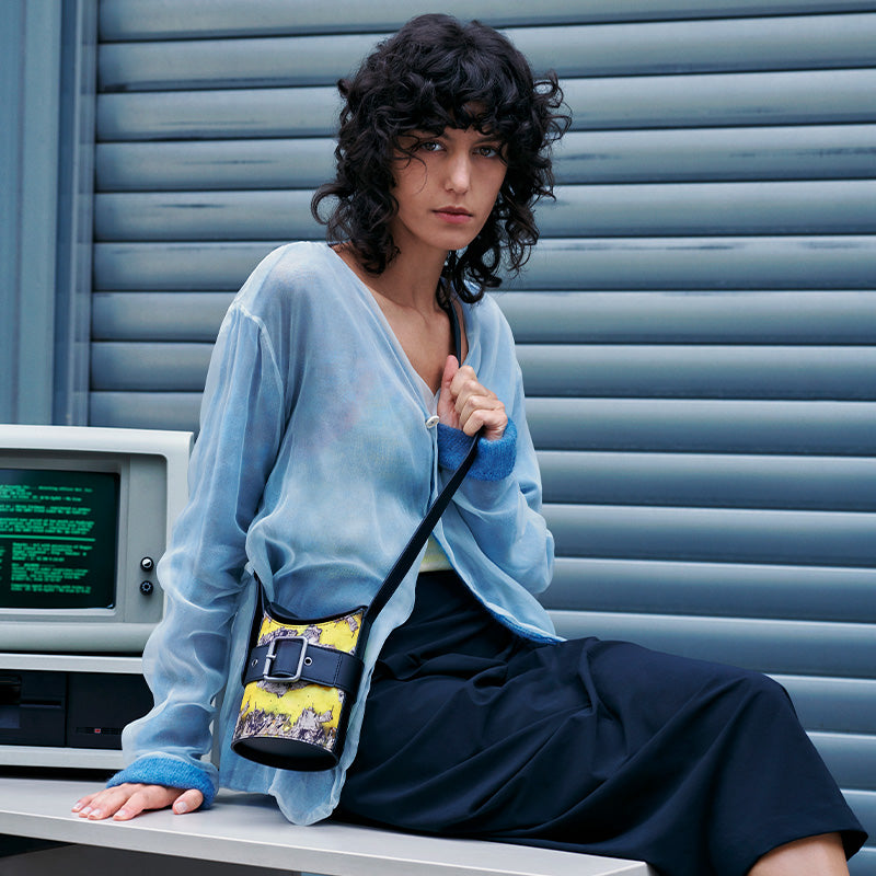 both x CHARLES & KEITH multi-color bucket bag worn by model with curly hair and blue top sitting near vintage computer